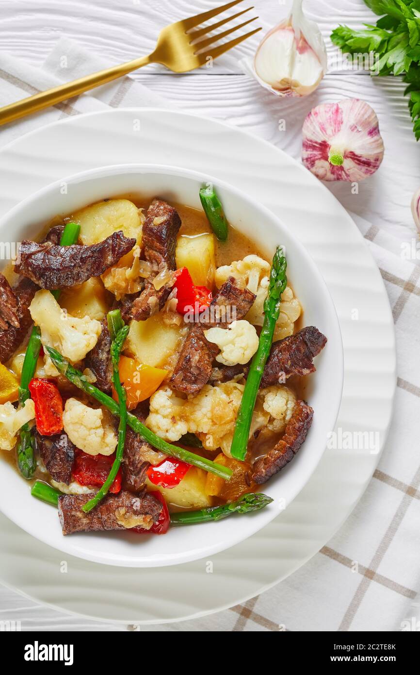 Nahaufnahme des Sommers Rinderstreifen Eintopf mit neuen Kartoffeln, Spargel und Blumenkohl in einer weißen Schüssel auf einem Holztisch, amerikanische Küche, vertikale Ansicht fr Stockfoto