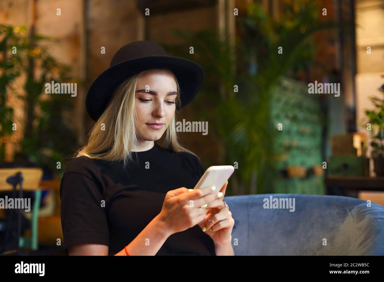 Blonde Mädchen in weißen Jeansrock, Bluse und schwarzen Hut, sitzen am Café-Tisch allein am Fenster, Blick auf das Handy Stockfoto