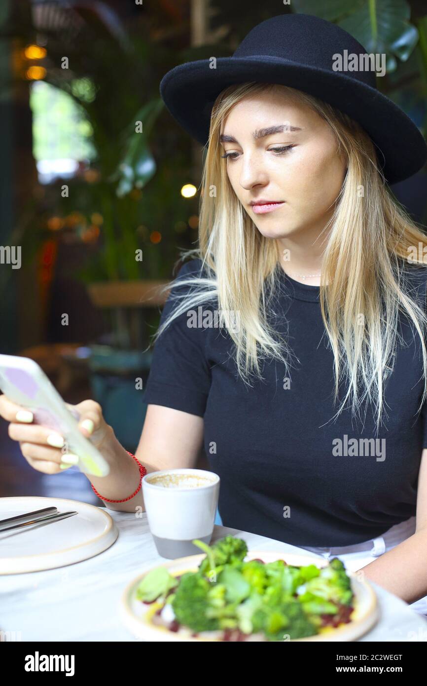 Attraktive niedliche kaukasische blonde Mädchen in weißen Jeans Rock, Bluse und schwarzen Hut Blick auf das Handy Stockfoto