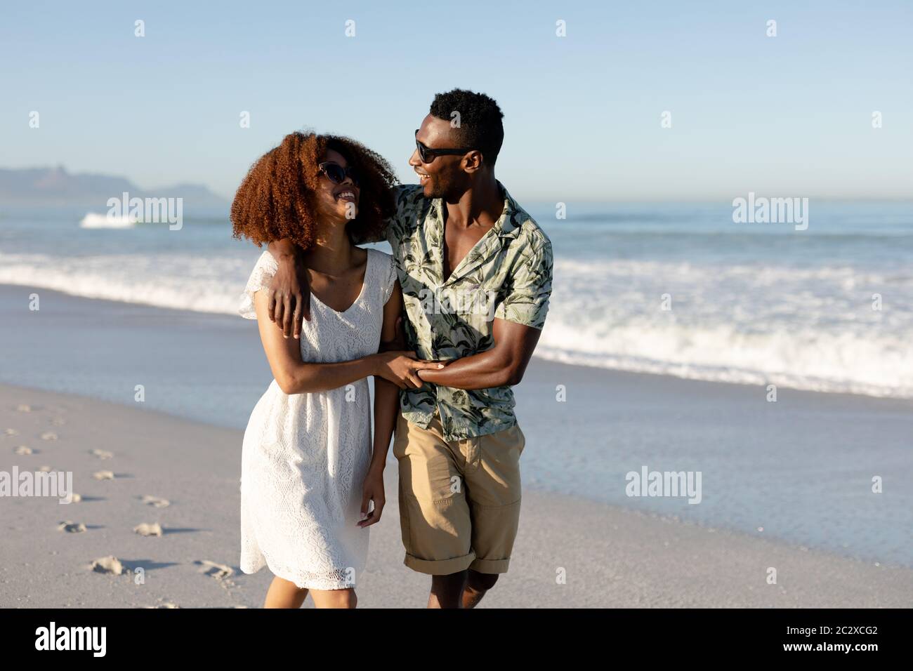 Ein gemischtes Rennpaar, das an einem sonnigen Tag am Strand spazierengeht und lacht Stockfoto