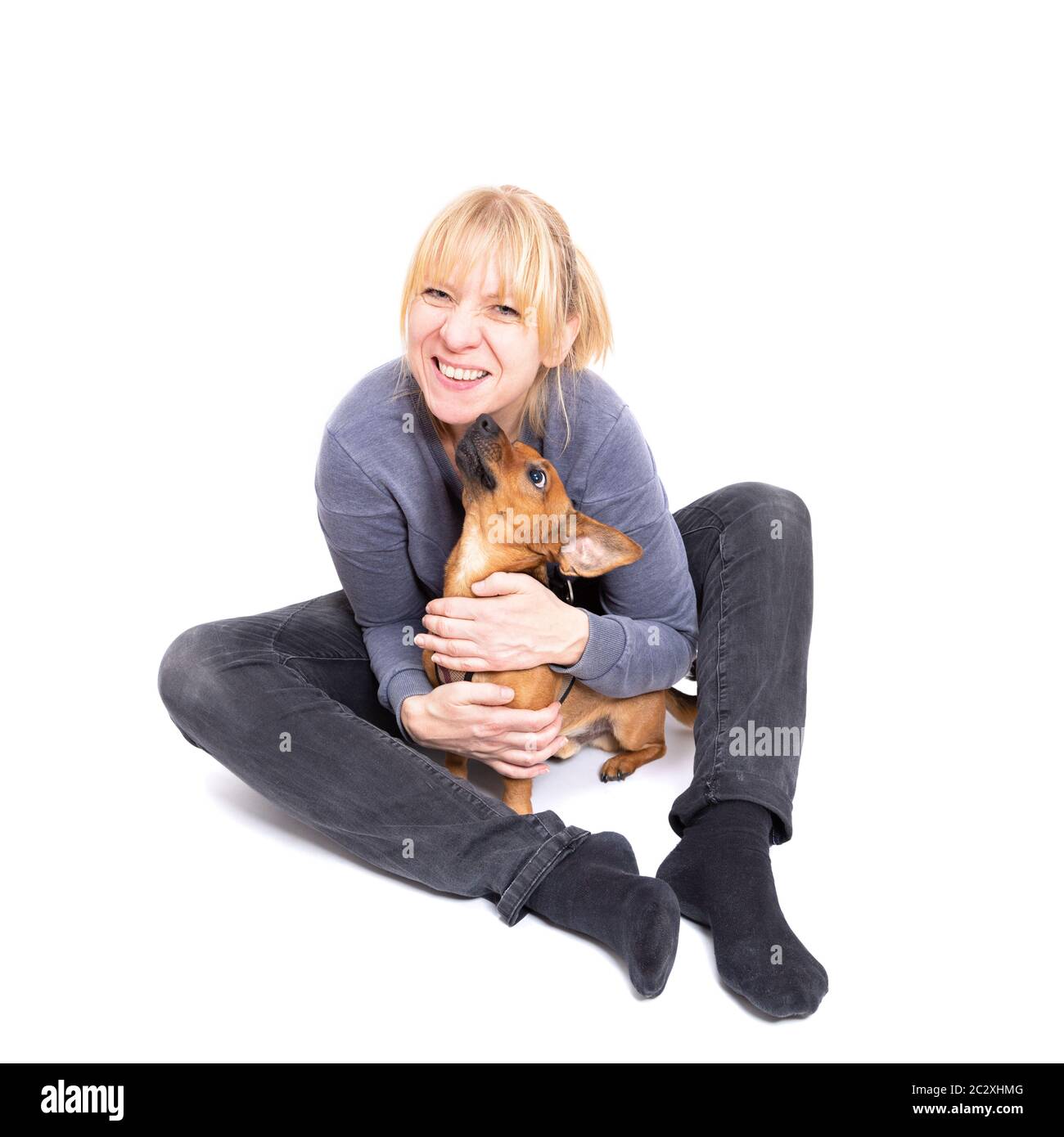 Blonde Frau sitzt auf dem Boden mit ihrem winzigen braunen Hund isoliert auf weißem Hintergrund im Studio. Kleine gemischte Rasse von Jack Stockfoto