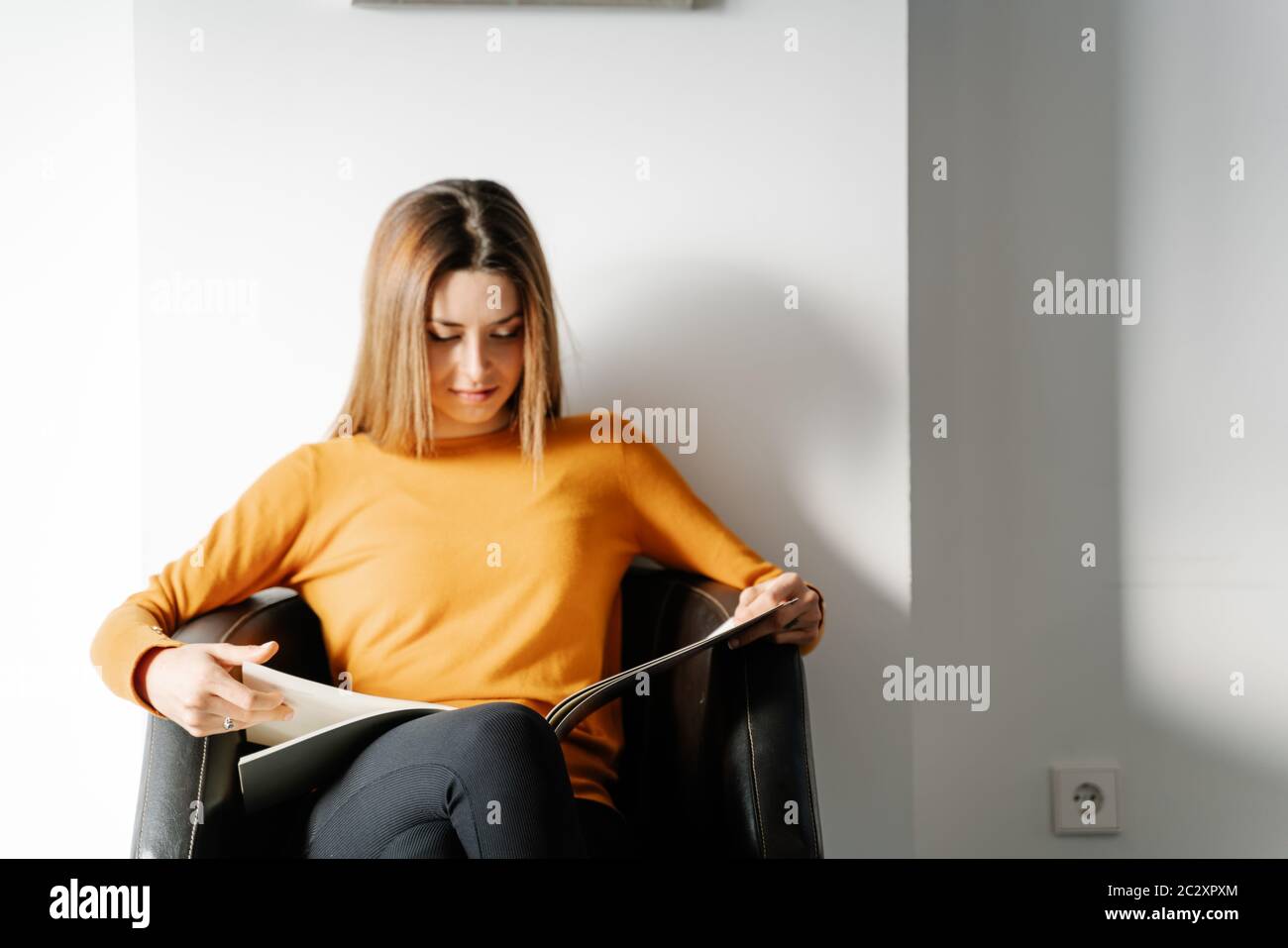Junge Frau in einem orangefarbenen Pullover wartet in einem Klinikbüro beim Lesen einer Zeitschrift Stockfoto