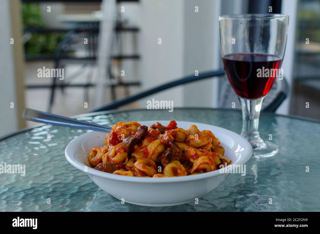 Nahaufnahme einer weißen Schüssel gefüllter Pasta ( Ravioli ) mit Tomatensauce und Pilzen mit einem Glas Rotwein auf einem Balkon Tisch im Freien Stockfoto