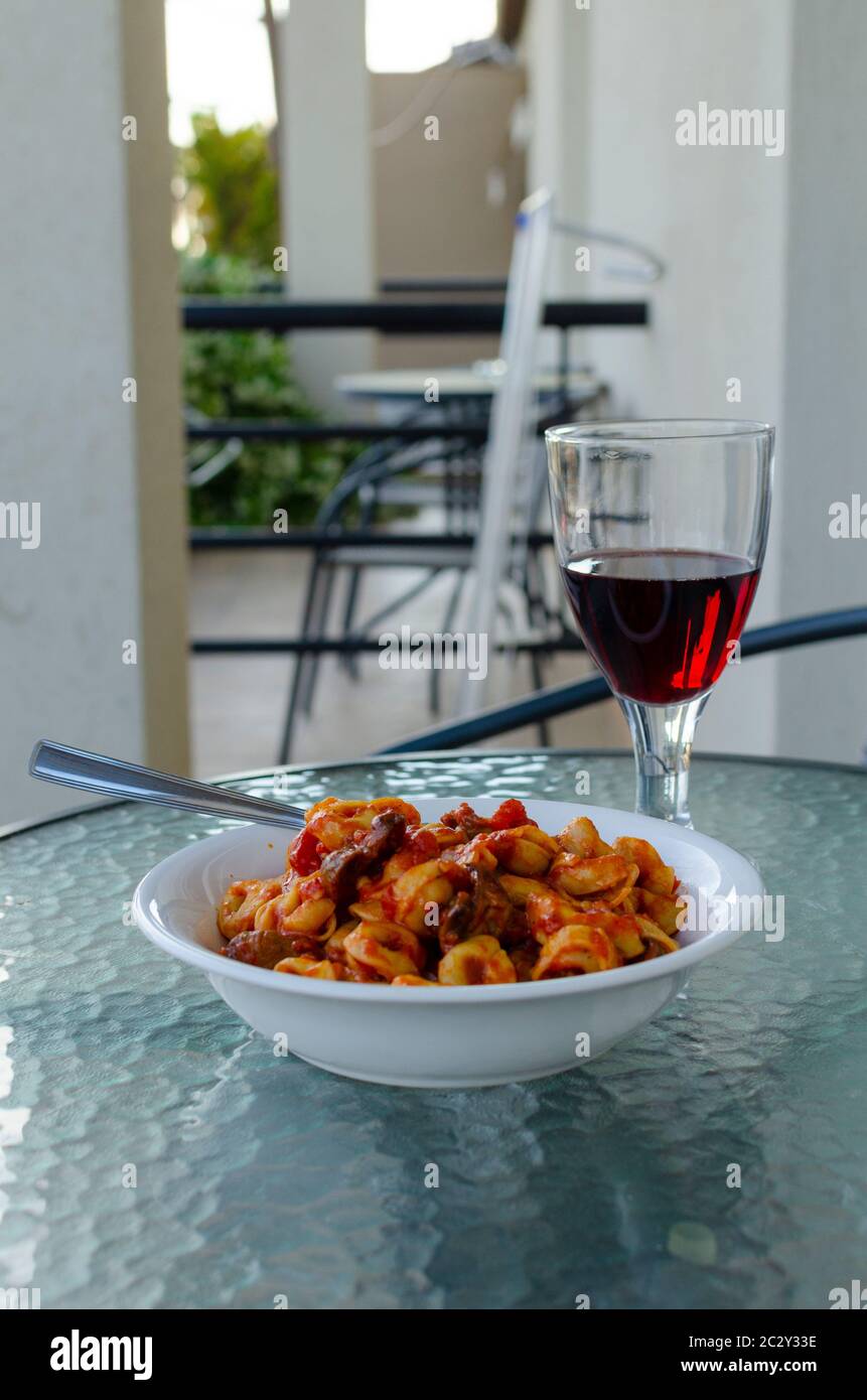 Nahaufnahme einer weißen Schüssel gefüllter Pasta ( Ravioli ) mit Tomatensauce und Pilzen mit einem Glas Rotwein auf einem Balkon Tisch im Freien Stockfoto