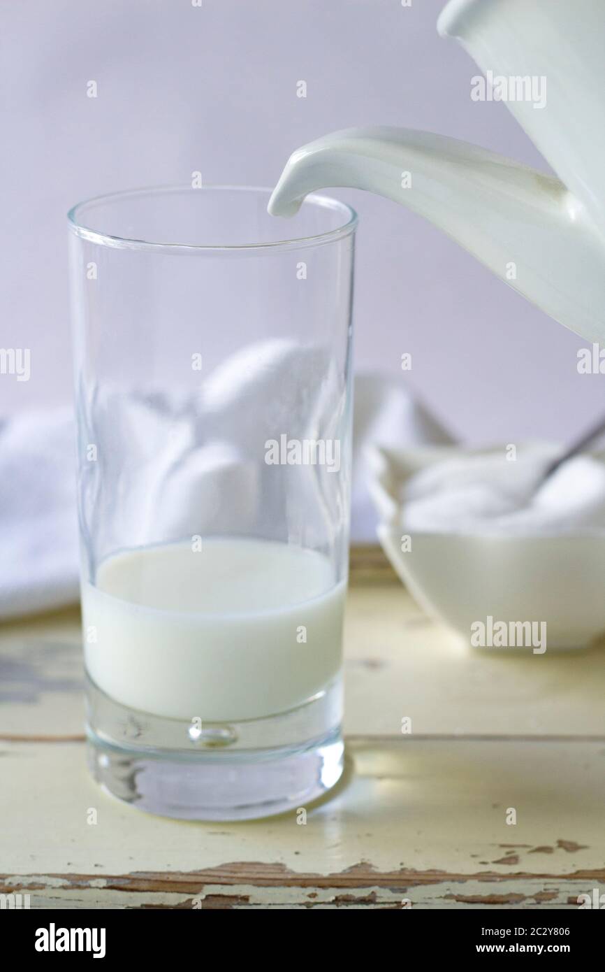 Detailaufnahme eines Milchkännchens, das ein Glas füllt, aber nicht zu füllen ist. Die köstliche Milch fällt nicht.Stillleben in weißem Ton. Im Hintergrund ein Zuckerbogen Stockfoto