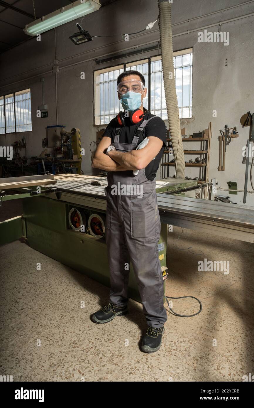 Arbeiter, der mit gekreuzten Armen steht, trägt Gesichtsmaske und Schutzkleidung und blickt in einer Fabrik lächelnd auf die Kamera. Langer Schuss, voller Körper. Stockfoto