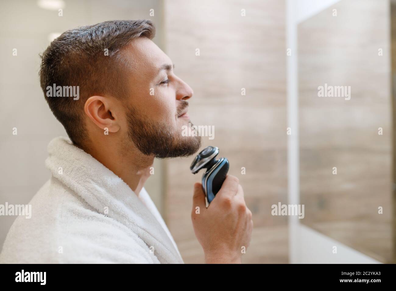 Mann am Spiegel rasiert seinen Bart mit einem elektrischen Rasierer im Bad,  Routine-Morgenhygiene. Männliche Person an der Spüle führt Haut und Körper  behandeln Stockfotografie - Alamy