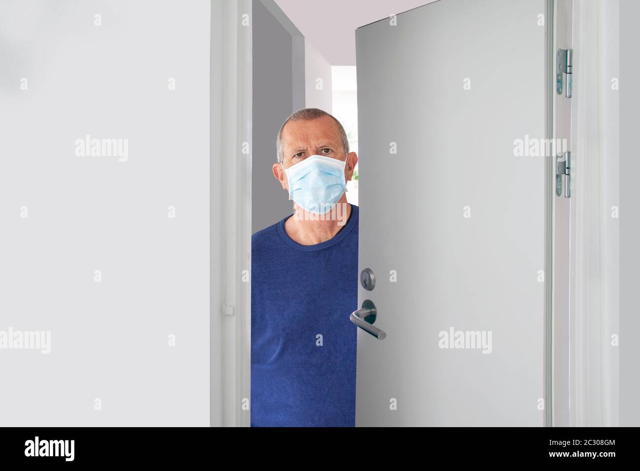 Älterer Mann mit medizinischer Gesichtsmaske, der in der Tür steht und halb offene Tür hat, die zu Hause oder im Krankenhaus schaut. Die Flurtür und das Innere sind alles Stockfoto