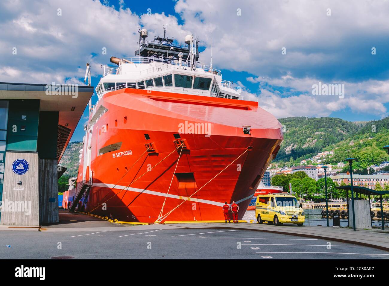 Zwei norwegische Sanitäter in roten Uniformen ruhen in der Nähe eines Krankenwagens, der in einem Hafen in der Nähe eines großen Schiffes geparkt ist. Thema Gesundheitswesen und Stockfoto