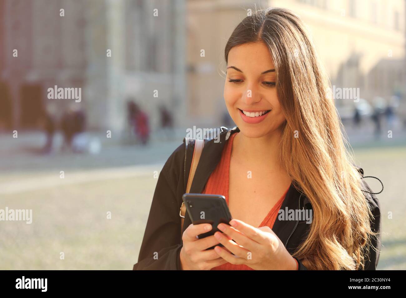 Fröhliches wunderschönes High-School-Mädchen lächelt und textet mit Smartphone im Freien bei Sonnenuntergang am Frühlingstag Stockfoto