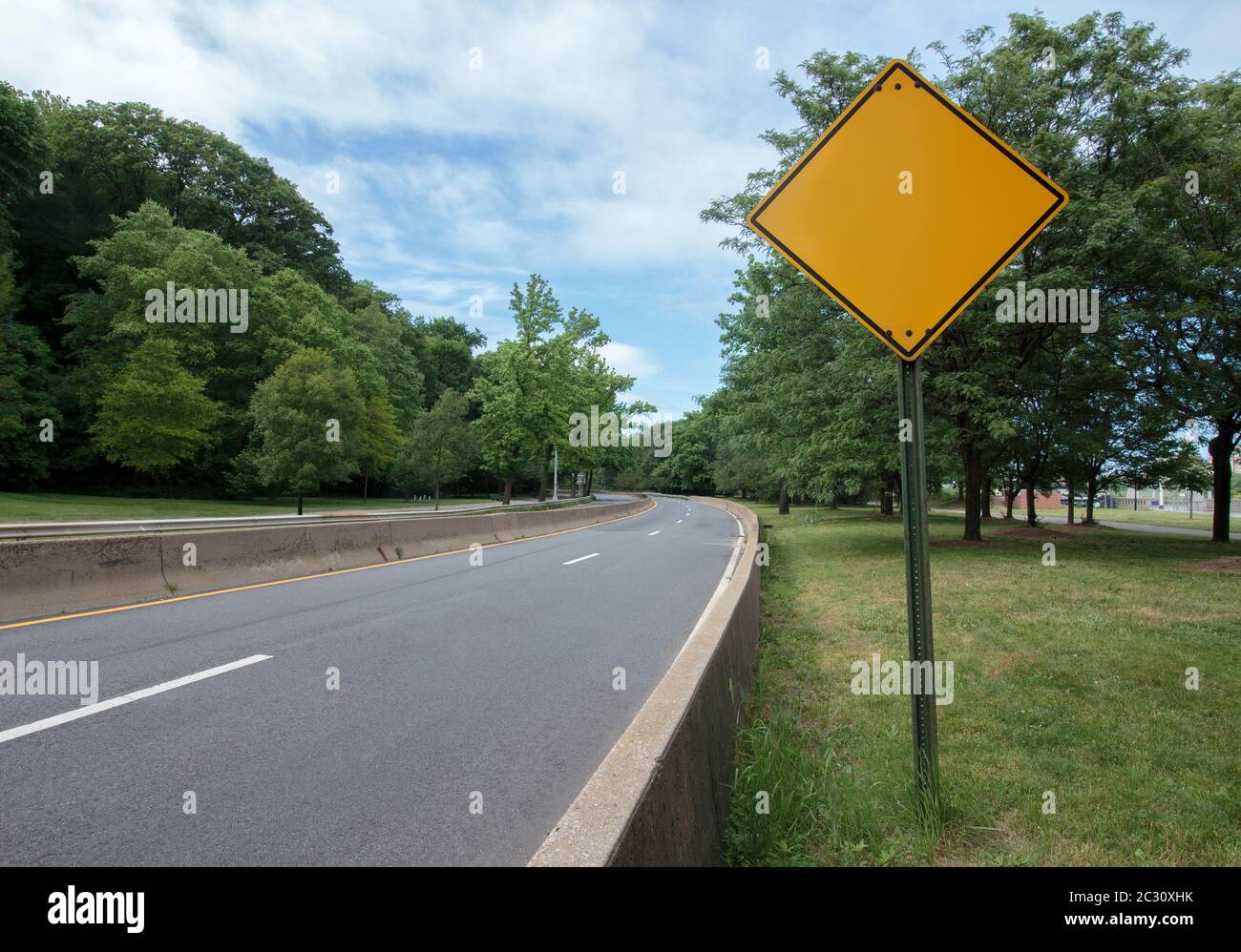Ein blank gelbes, diamand geformtes Schild an der Seite einer leeren Autobahn mit Bäumen auf beiden Seiten Stockfoto