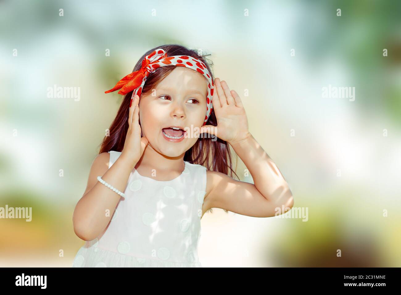 Happy Kid Mädchen Hand zu Ohr Lauschangriffe versuchen, ein Geplauder zu hören. Nahaufnahme Porträt des kaukasischen Kind Modell, rot weiß Stirnband isoliert grünen Baum pa Stockfoto