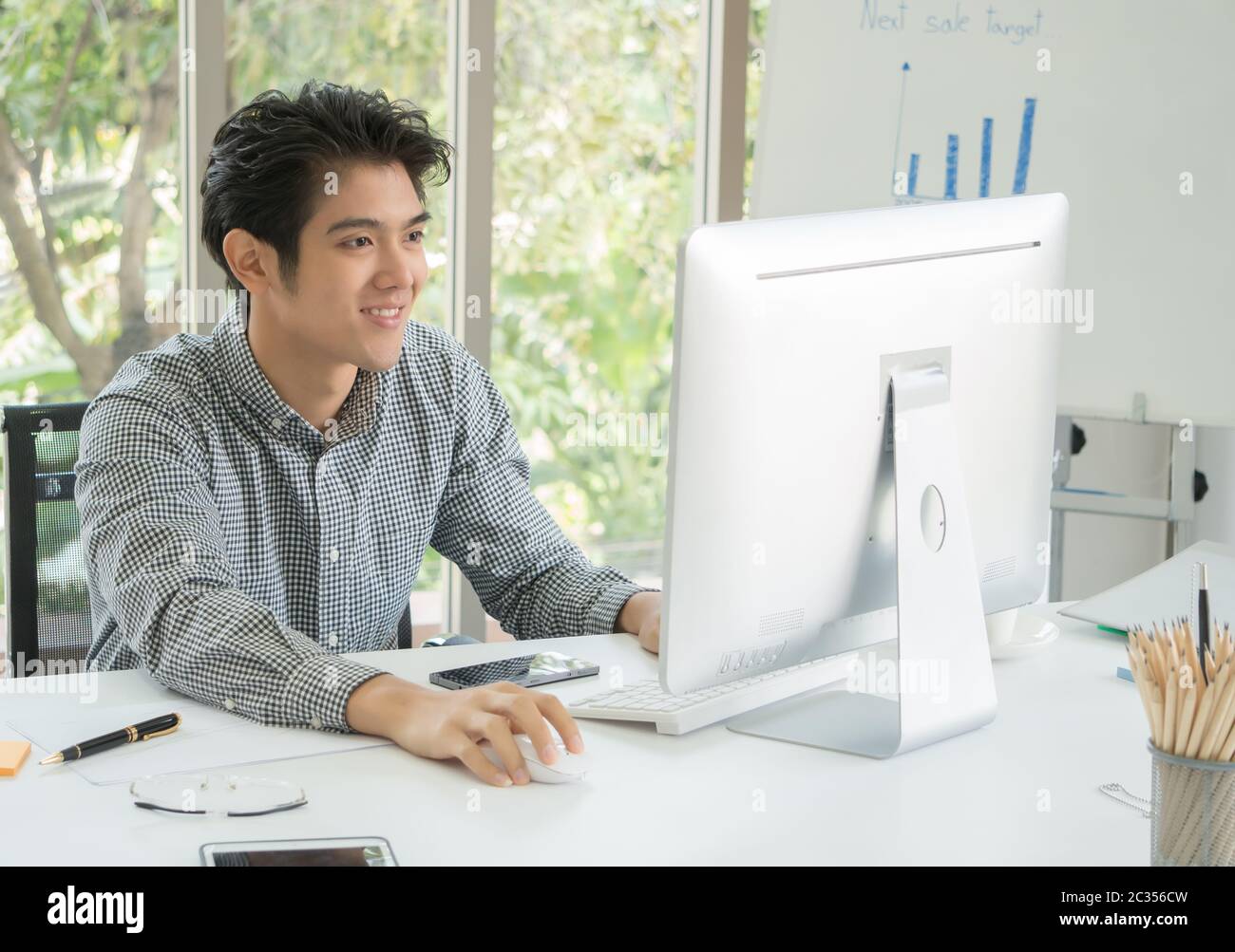 Der stattliche Asiat im Alter von 25 bis 35 Jahren sitzt am Schreibtisch und lächelt fröhlich. Er ist der Leiter des Marketings. Er überwacht den Umsatz auf dem Computerbildschirm. Stockfoto