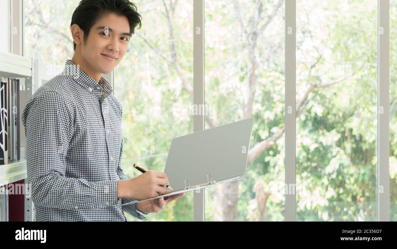 Ein 25-35-jähriger asiatischer Geschäftsmann lächelte glücklich im weißen Büro. Er überprüfte die Dokumente, um die Handelsdokumente zu unterzeichnen. Stockfoto