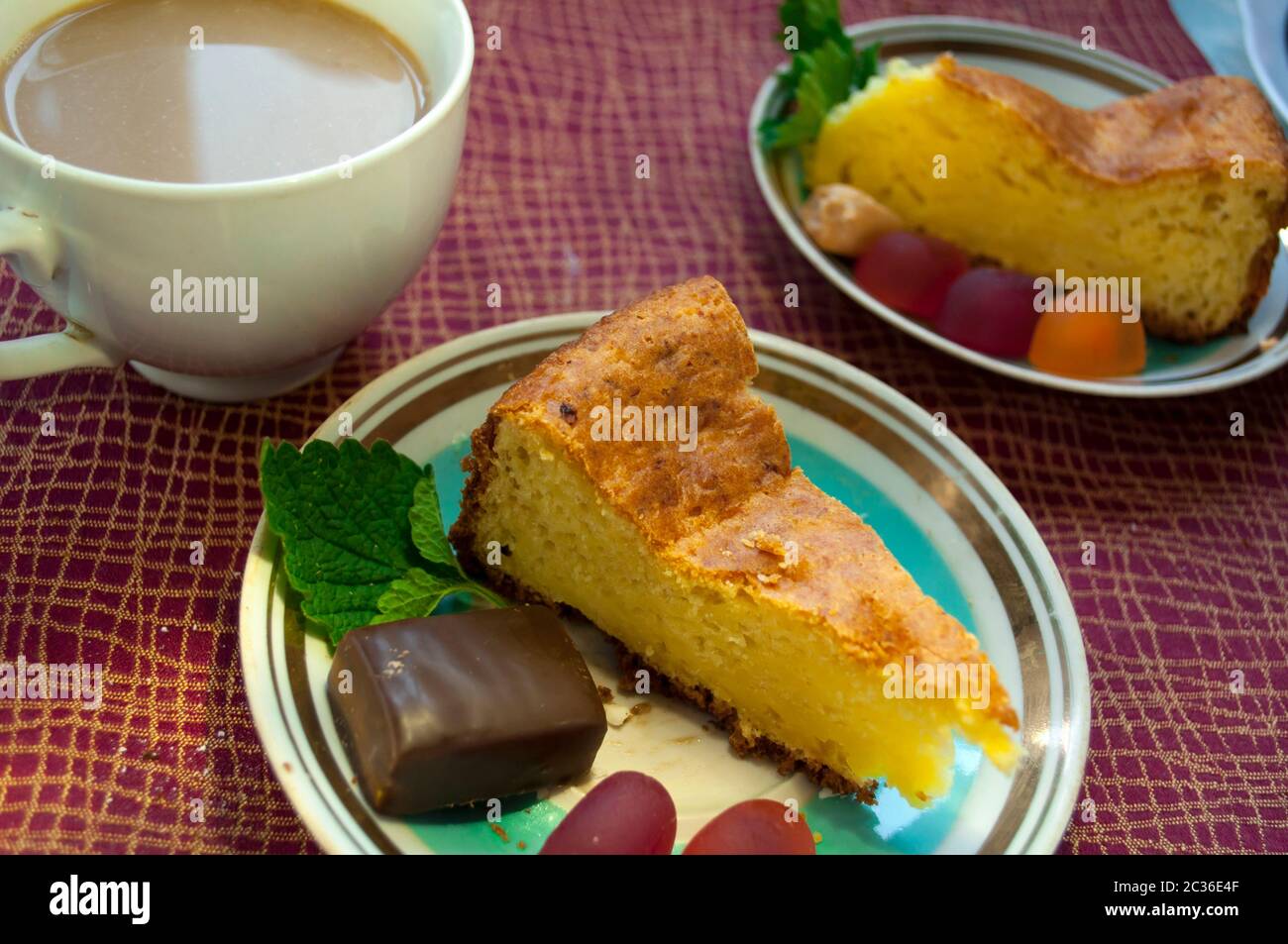 Kuchen und Kaffee mit Milch auf einer Tischdecke burgunderrot Farbe. Ein Stück Kuchen mit Gelee Bonbons und Schokolade Bondi wird auf einer Untertasse genommen Stockfoto