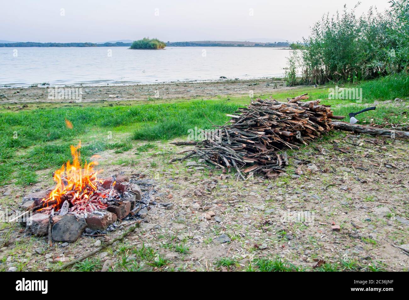 Kleines Lagerfeuer mit Flammen neben einem See mit Holzstapel an der Seite. Stockfoto