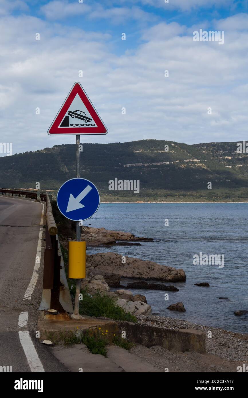 Straße den Hügel hinauf am Ufer des Mittelmeers mit zwei Verkehrsschildern. Ruhiges Wasser des Meeres. Berge im Hintergrund. Blauer Himmel mit Inten Stockfoto