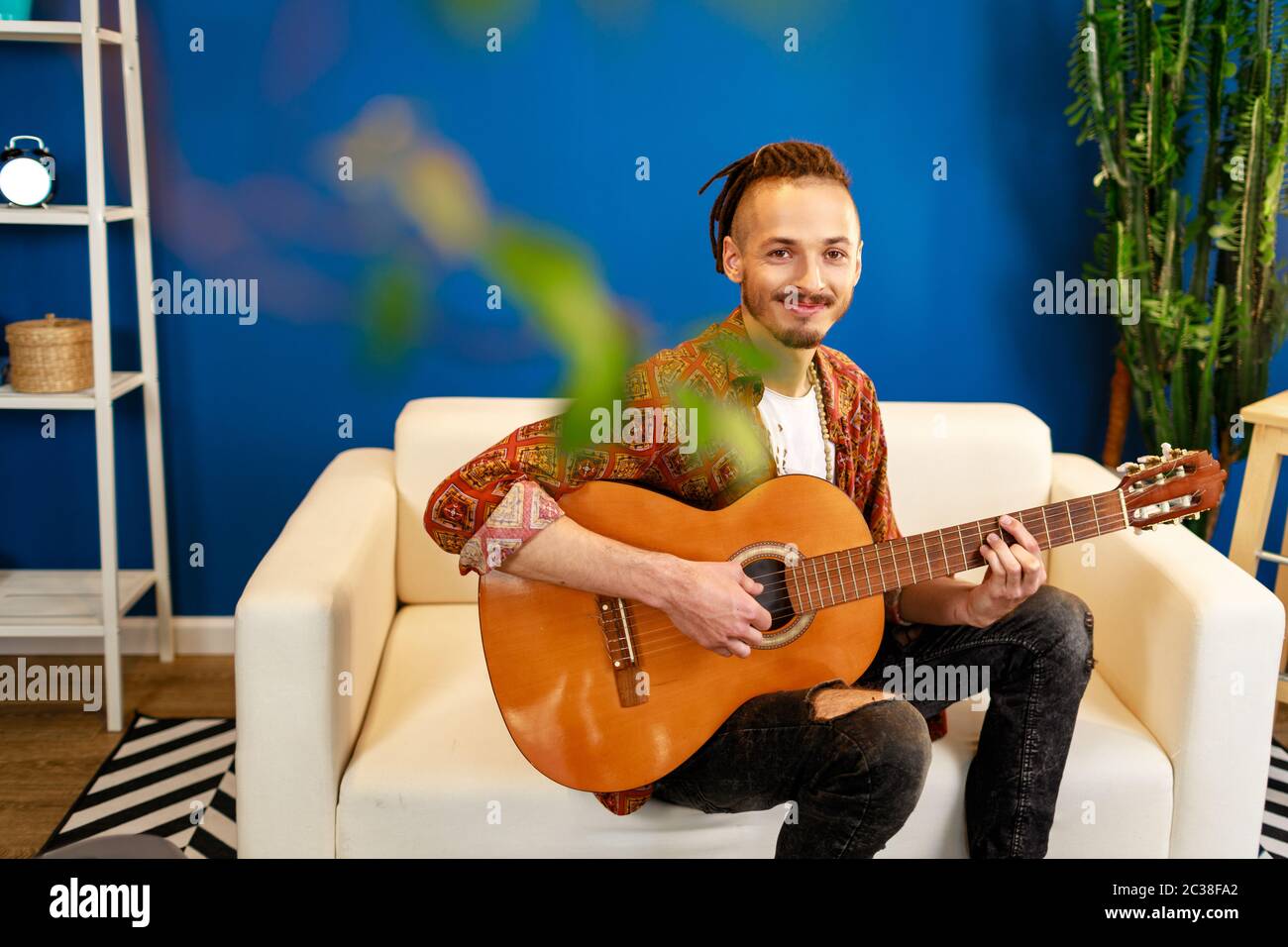 Junger kaukasischer Mann mit Dreadlocks auf dem Sofa sitzend und akustische Gitarre spielend Stockfoto