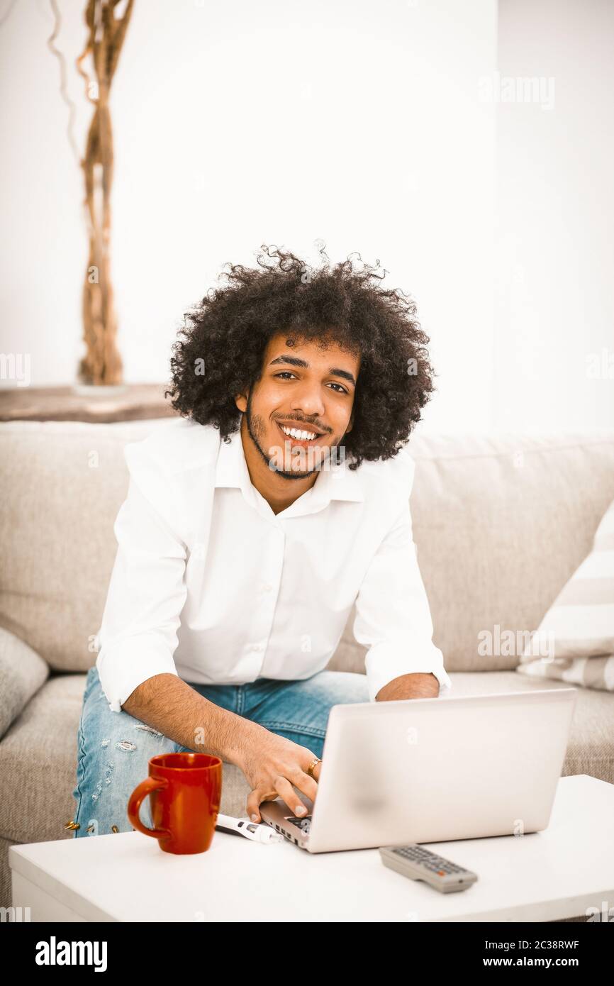 Arabischer Student arbeitet Laptop am Couchtisch mit roter Tasse darauf. Schöner Mann lächelt siiting auf Sofa zu Hause. Home Education Konzept. Getöntes Bild Stockfoto