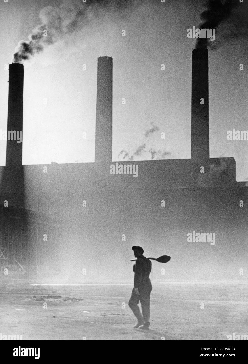 Ein Arbeiter geht auf dem Industriegelände. Im Hintergrund die Schornsteine der Fabrik. Stockfoto