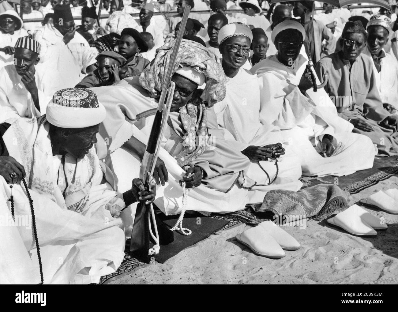 Muslimische Männer sitzen auf Gebetsteppichen beim Ramadan-Fest in Medina, einem Stadtteil von Dakar. Ein Mann hält ein Gewehr. Die anderen Männer tragen Gebetsketten. Stockfoto