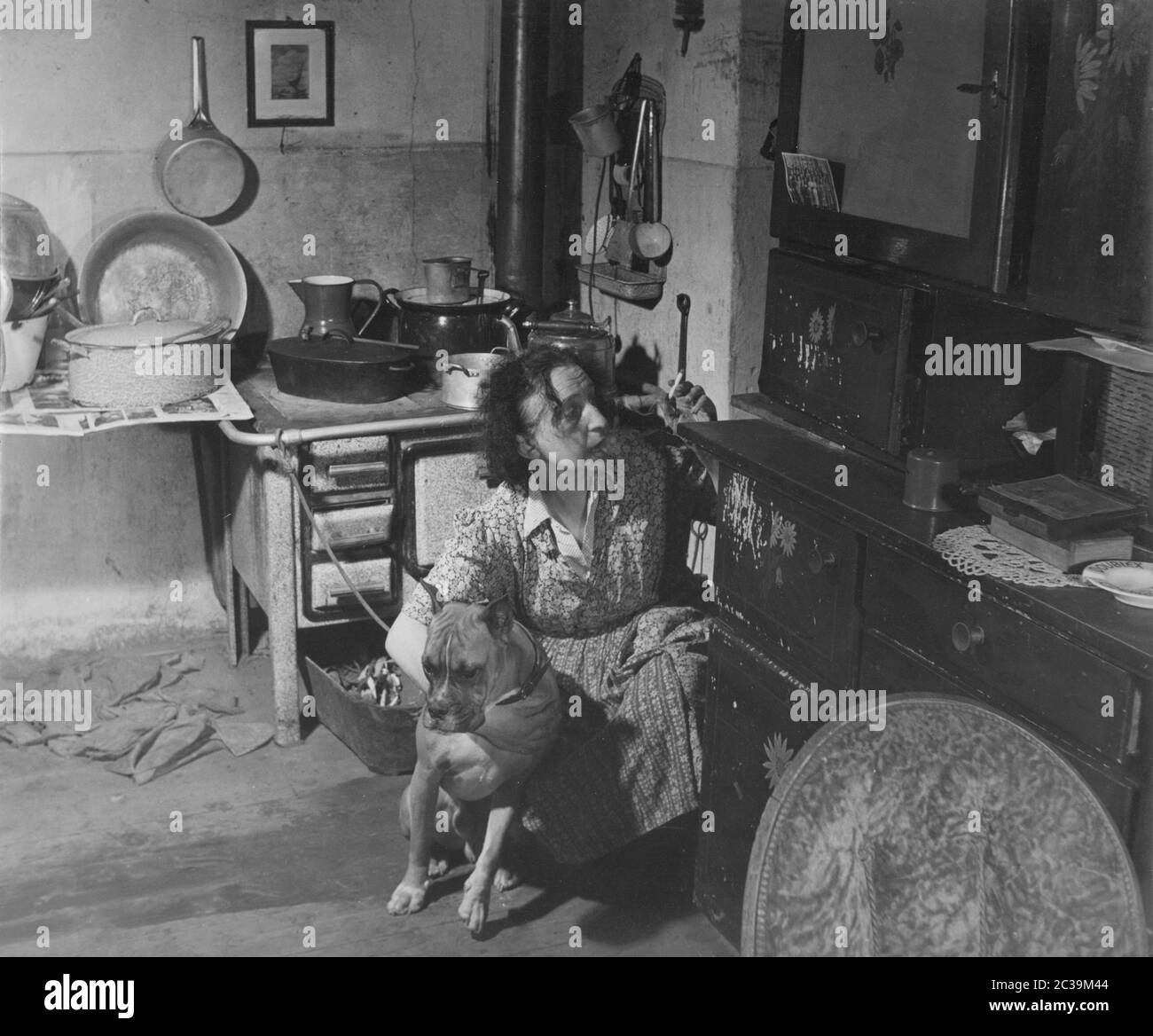 Eine rauchende Frau in der Schürze einer traditionellen Hausfrau in einer altmodischen Küche hält eine Bulldogge in ihren Armen. Undatierte Aufnahme. Stockfoto