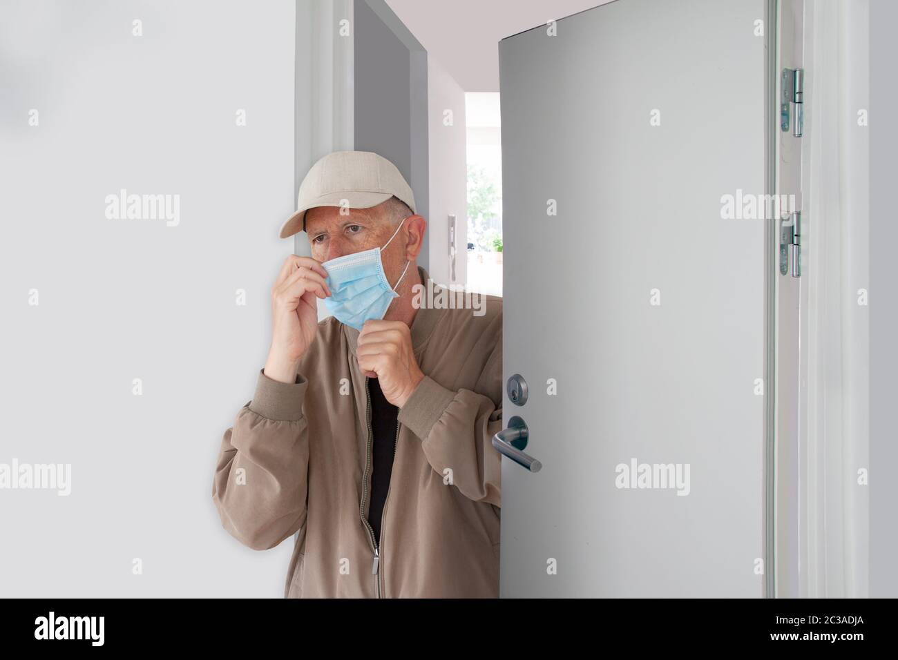 Mann mit Kappe, der seine schützende medizinische Maske anlegt, als er das Haus verlässt. Er geht eine halb geöffnete Tür in einem Gebäude hinaus. Coronavirus Covid-19 Pandemie machen Stockfoto