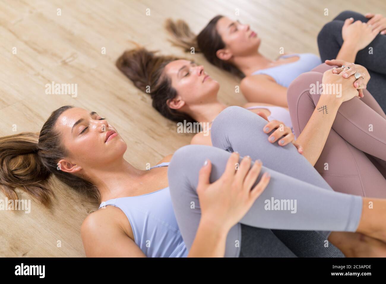 Gruppe von drei jungen sportlich attraktiven Frauen im Yoga-Studio, auf dem Boden liegend, dehnend und entspannend nach dem Training. Gesunde, aktive Lebensweise, Stockfoto