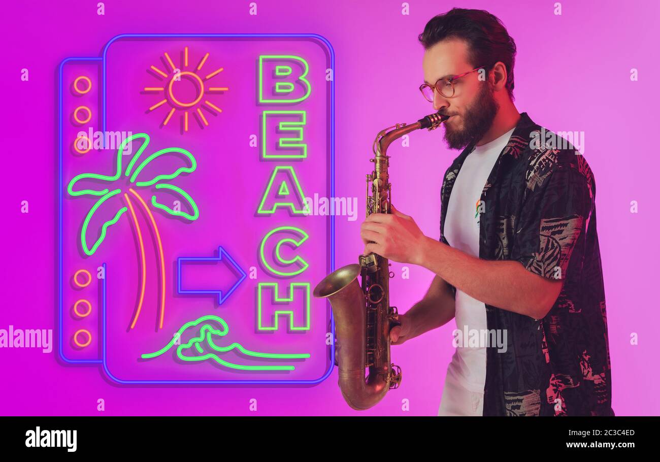 Junger Jazzmusiker spielt Saxophon auf gradient Studio Hintergrund in Neonlicht mit Schild STRAND. Konzept der Musik, Hobby, Festival. Fröhlich inspirierter Künstler, Flyer. Sommer, Verkauf, Urlaub. Stockfoto