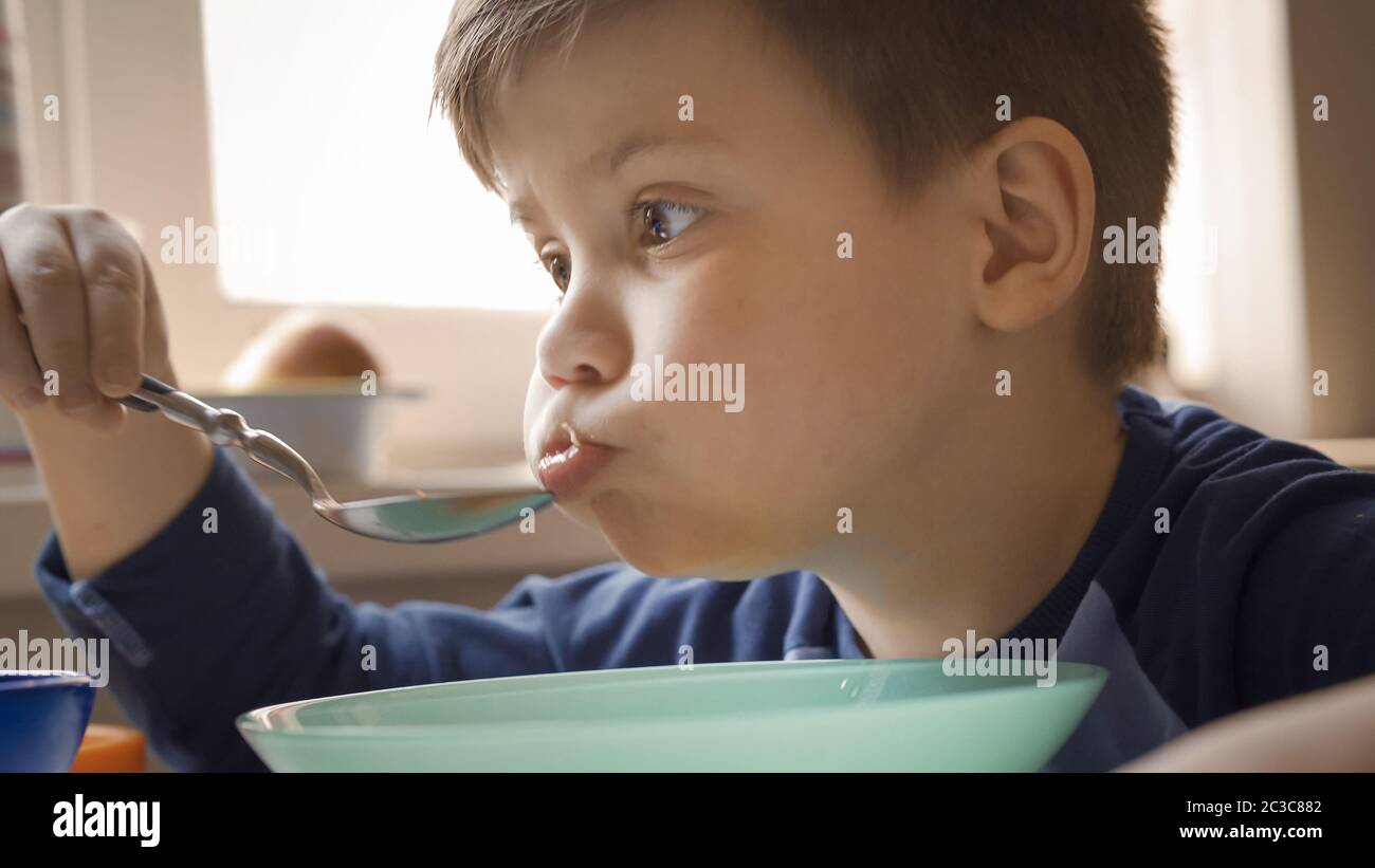 Vorschulkinder essen Suppe selbst. Junge mit vollen Wangen isst am heimischen Küchentisch sitzen. Gesundes Lebensmittelkonzept. Nahaufnahme Stockfoto