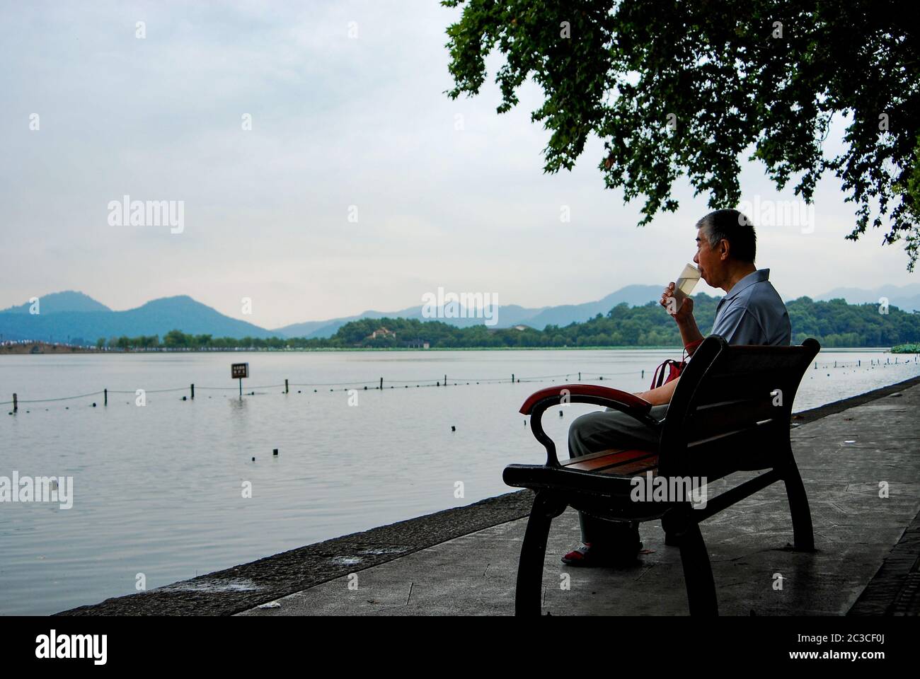 Ein älterer Mann, der an der Bank sitzt und seinen Tee aus einer wiederverwendbaren Plastikflasche trinkt, während er an bewölktem Tag die Xihu, das Wasser des Westsees, betrachtet Stockfoto