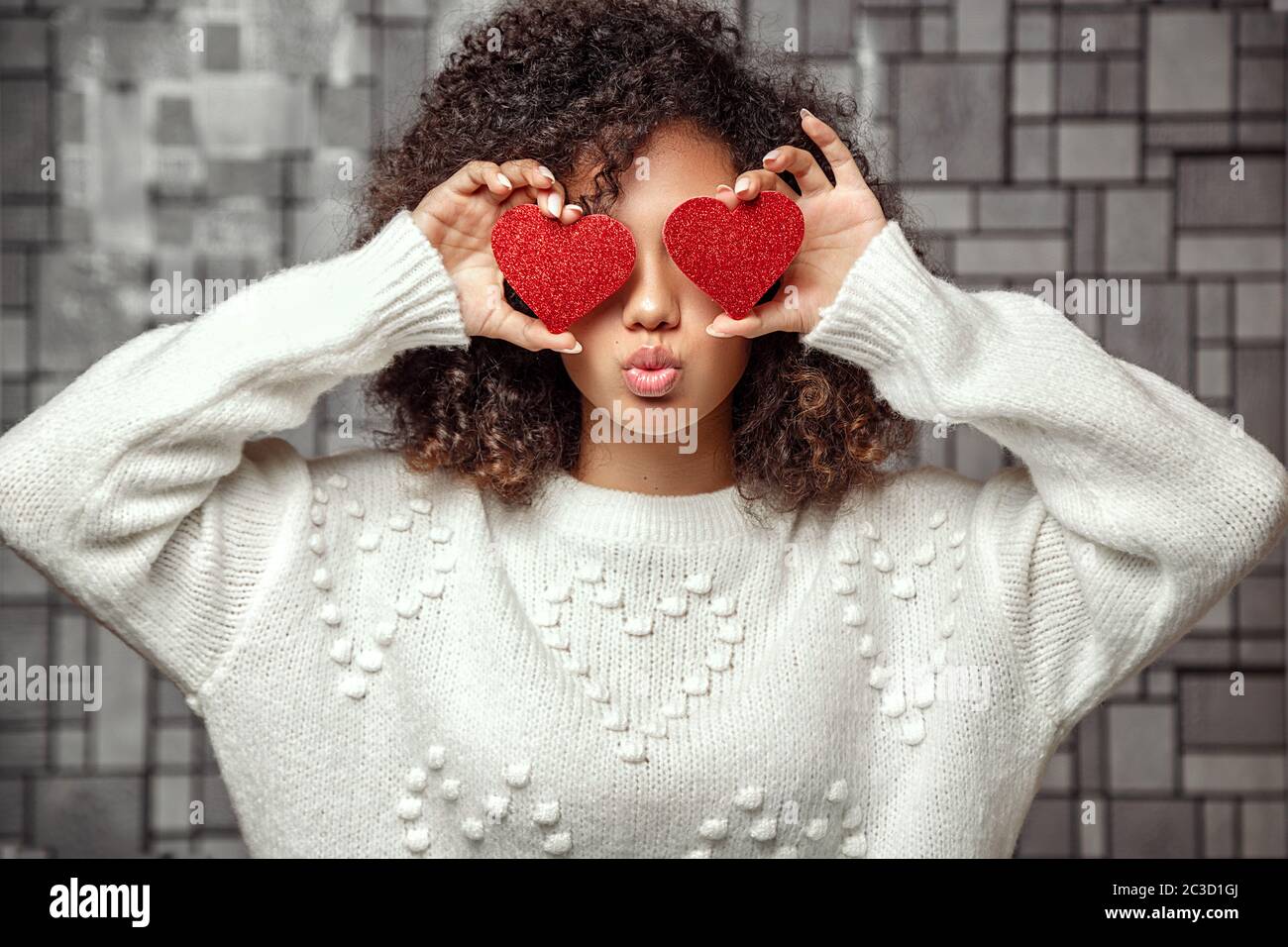 Ein junges afroamerikanisches Mädchen mit lockigen Haaren in einem weißen Pullover hält zwei Herzen in den Händen. Schließt die Augen mit Herzen und macht Stockfoto