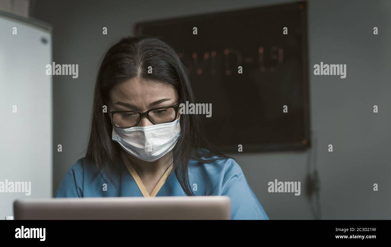 Müde Ärztin, die mit Laptop arbeitet. Hübsche asiatische Frau in Maske und Brille Blick auf Computer-Bildschirm. Nahaufnahme. Überarbeitungskonzept. Getönte Stockfoto