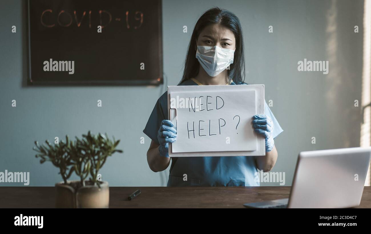 Seriöse Arzt bietet Hilfe an. Asiatische Frau in Schutzuniform mit langen Haaren schaut auf Kamera stehen in ihrem medizinischen Büro. Getöntes Bild Stockfoto