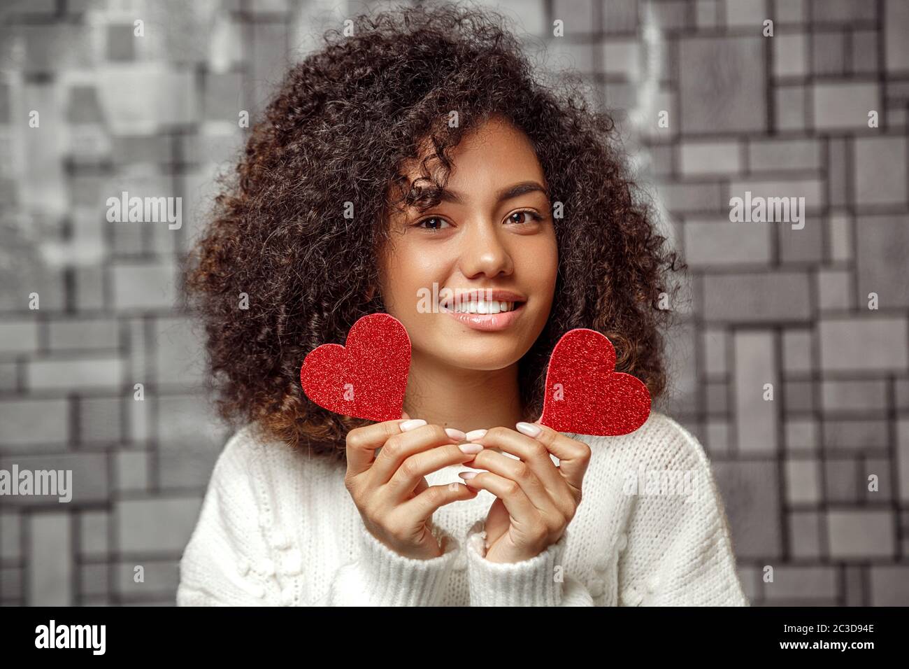Nahaufnahme Porträt eines jungen lockigen afroamerikanischen Mädchen in einem weißen Pullover mit zwei Papier Herzen. Flache Tiefe der Stockfoto