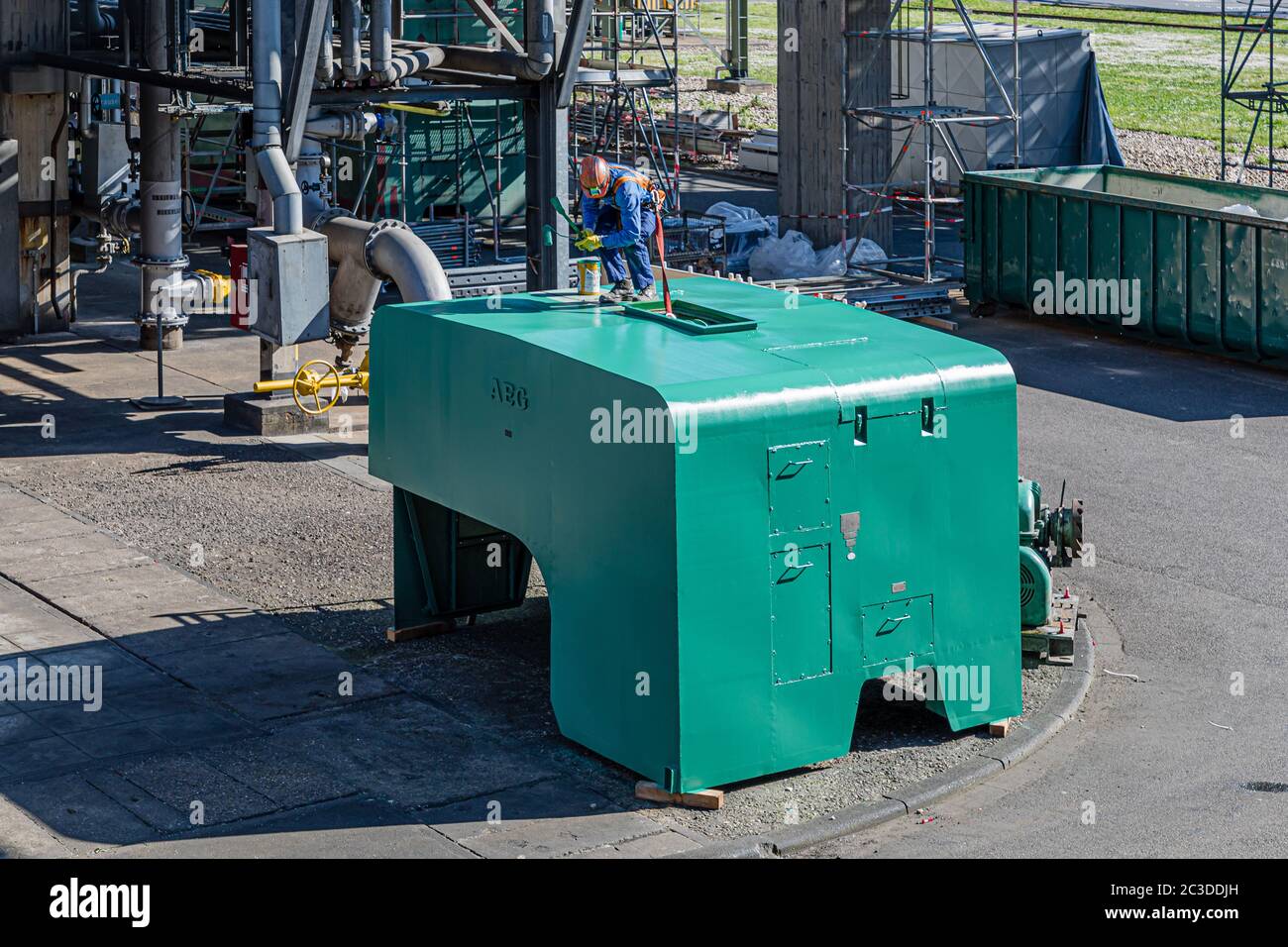 Ein Arbeiter bemalt die Verkleidung eines Kompressors in einer Chemiefabrik, die mit einem Sicherheitsgurt und einem Fallschutz gesichert ist. Stockfoto