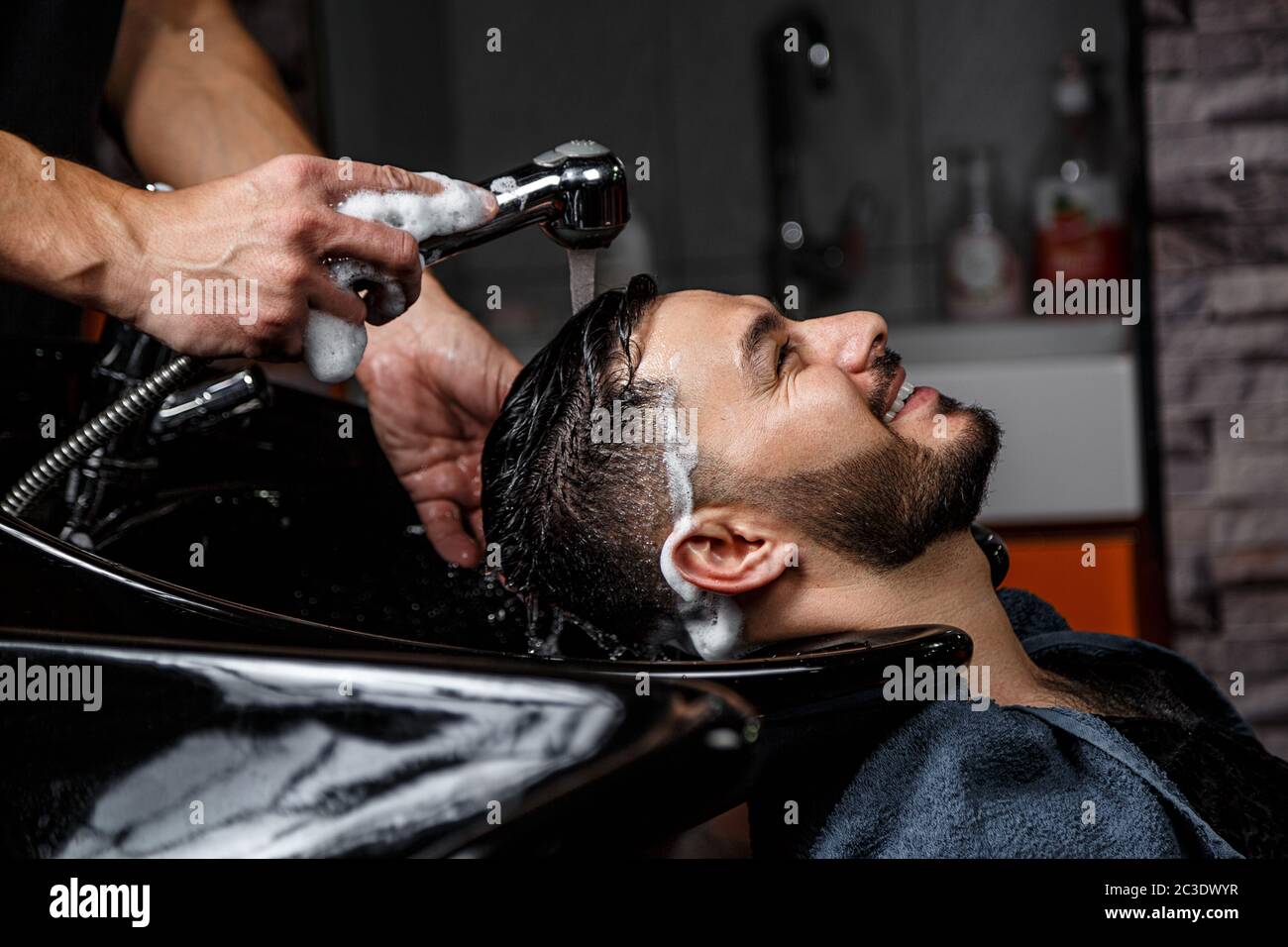 Ein junger dunkelhaariger Kerl mit indischem oder asiatischem Auftritt in einem Barber-Shop auf einem schwarzen Stuhl. Der Barber wäscht seinen Kopf mit Shampoo Stockfoto