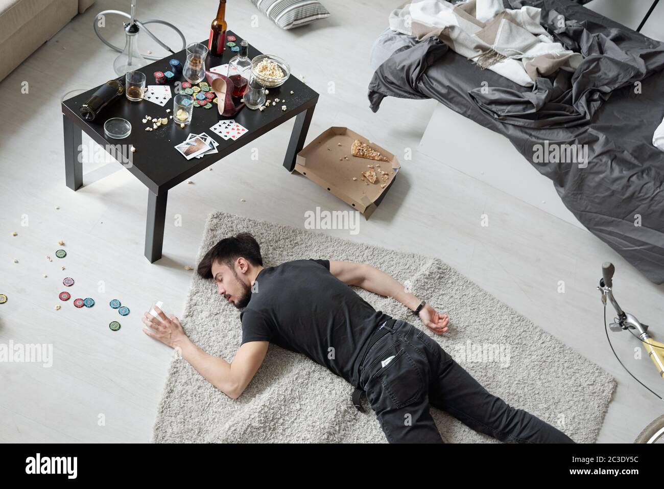 Oben Blick auf betrunkenen Mann in schwarzem Outfit schlafen auf dem Teppich unter Krümel und Poker-Chips nach der Party Stockfoto