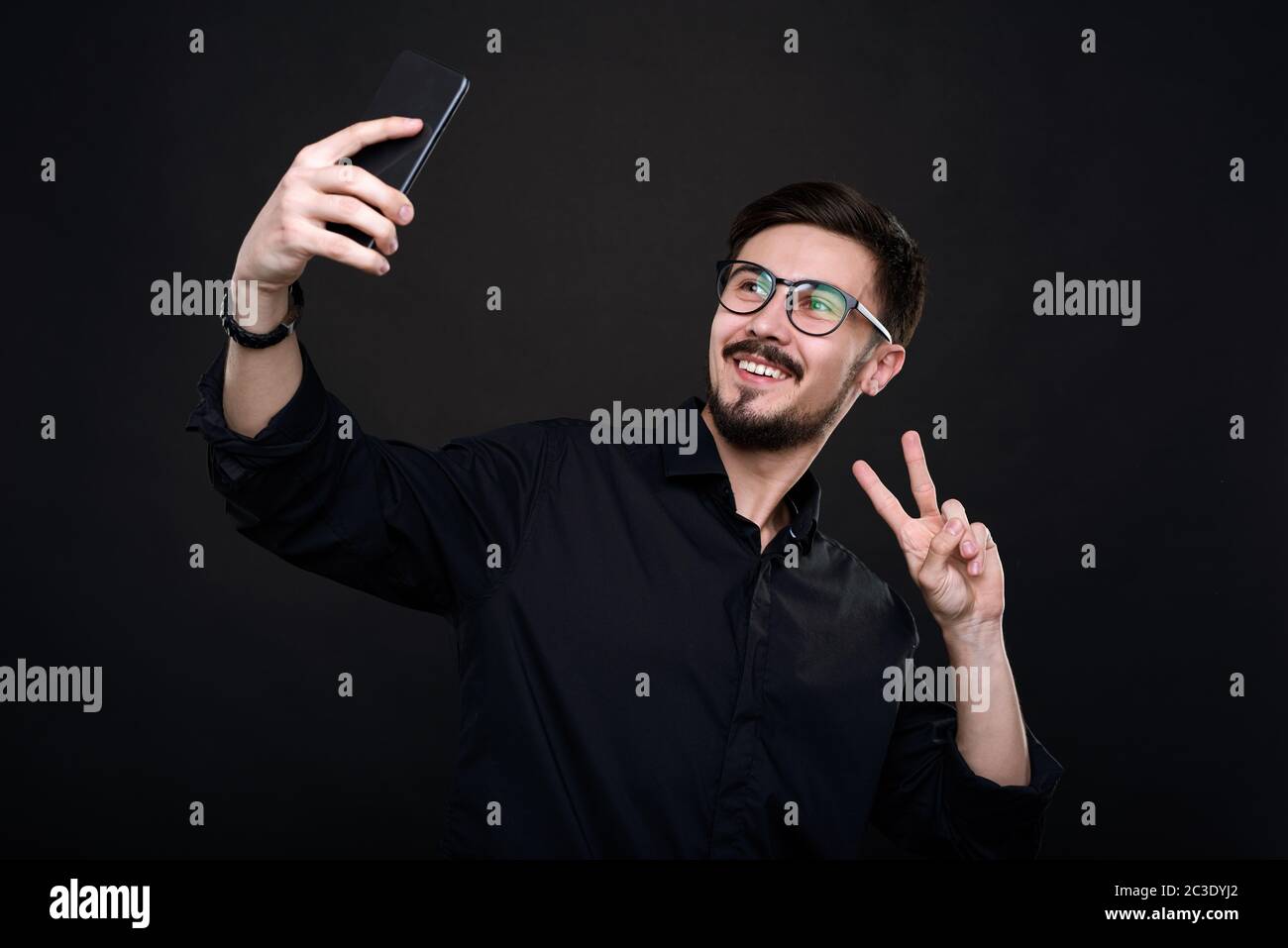 Lächelnder, hübscher junger Mann in einer Brille, der ein Friedenszeichen zeigt, während er Selfie auf dem Smartphone nimmt Stockfoto