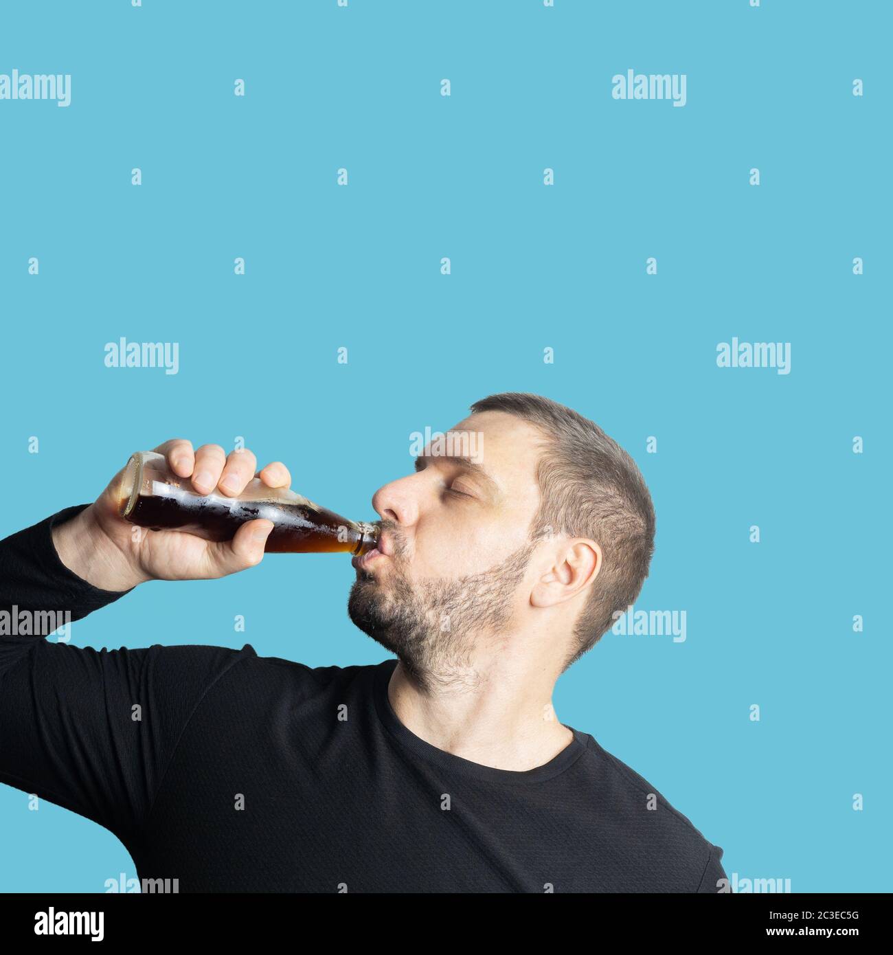 Dunkelhaariger bärtiger Mann in dunkler Kleidung trinkt mit geschlossenen Augen ein dunkles Getränk aus einer Flasche Stockfoto