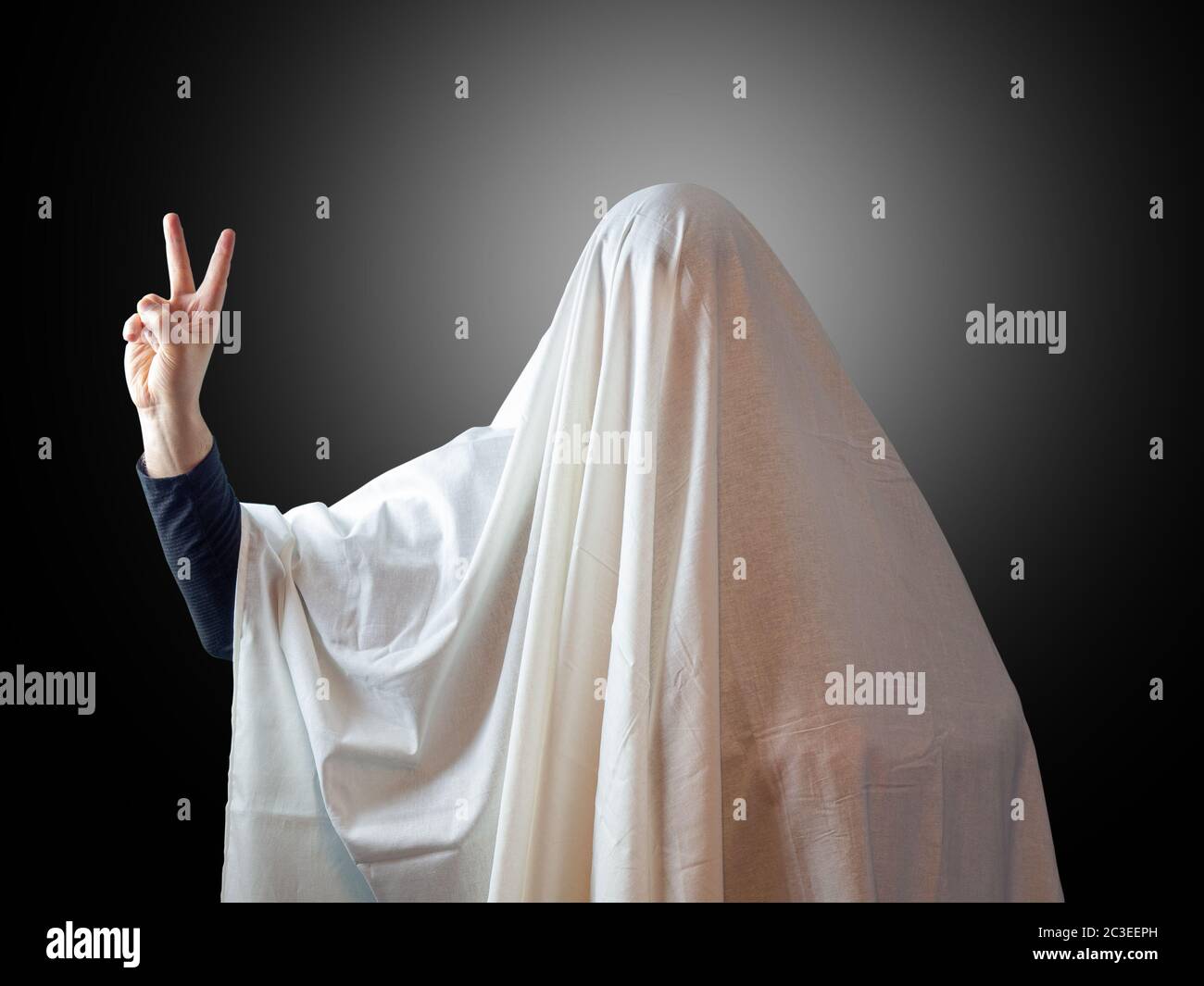 Ein Mann, der mit einem Blatt wie ein Geist bedeckt ist, zeigt zwei Finger, die den Sieg auf einem dunklen Hintergrund mit Hintergrundbeleuchtung im Hintergrund anzeigen Stockfoto