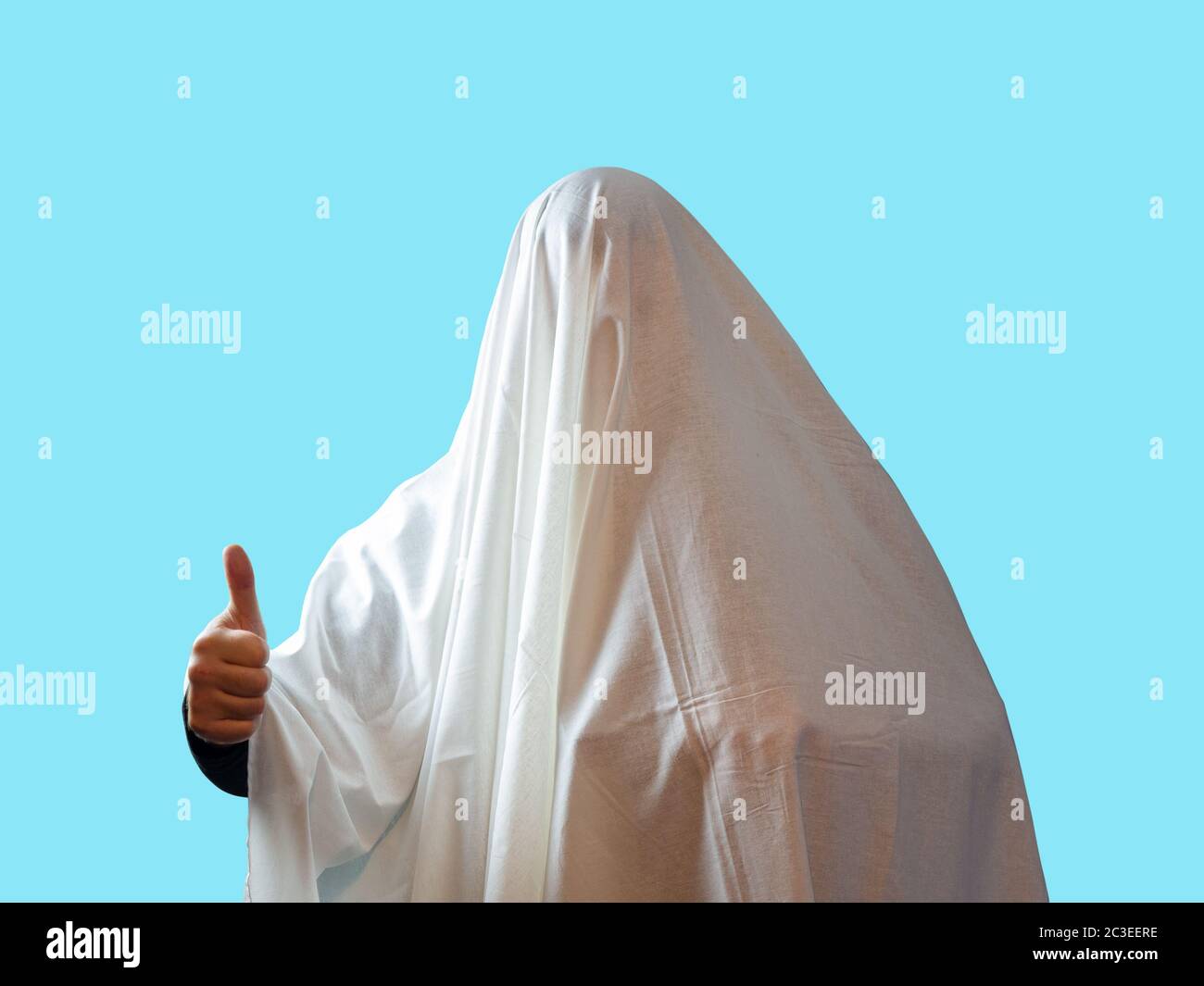 Ein Mann, der mit einem weißen Blatt wie ein Geist bedeckt ist, zeigt einen Daumen auf blauem Hintergrund Stockfoto