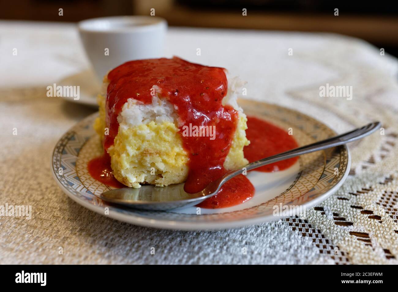 Stück Käsekuchen mit Erdbeerdecke auf einem Teller mit einem Löffel, auf einer weißen Tischdecke mit einer Tasse im Hintergrund Stockfoto