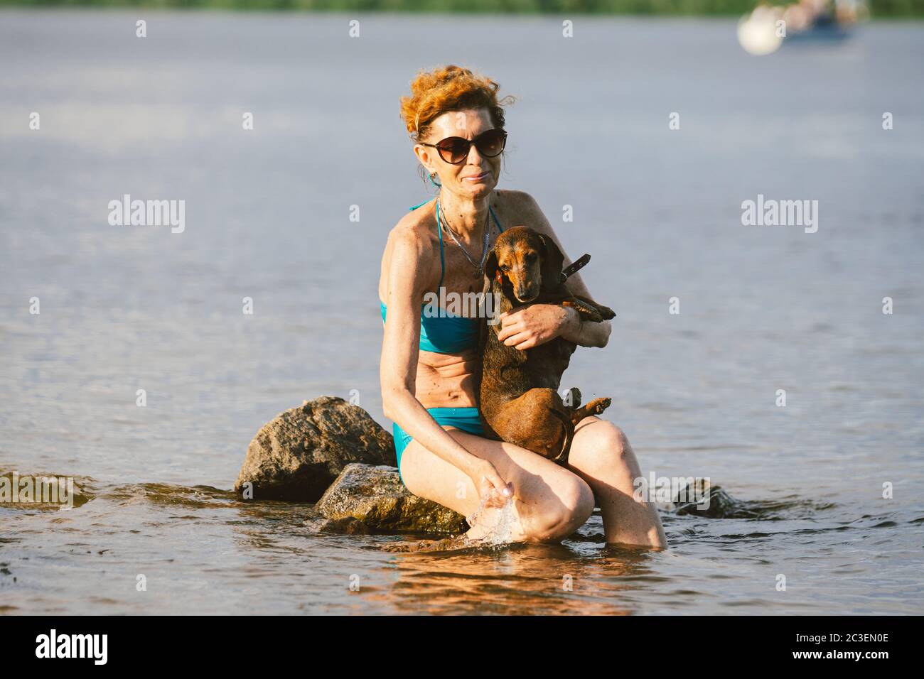 Reife Frau genießen Tag am Strand mit Hund. Die Besitzerin, Hündin im Badeanzug und kleiner, brauner Dackel-Hund, verbringt ihre Sommerzeit in Kälte Stockfoto