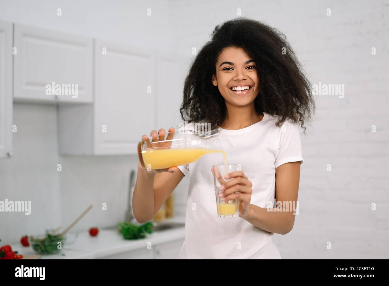 Junge schöne afroamerikanische Frau hält Glas mit einem Orangensaft, Blick auf die Kamera und lächelt. Gesundes Lifestyle-Konzept Stockfoto