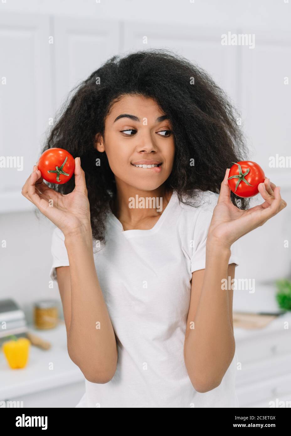 Schöne afroamerikanische Frau Wahl zwischen zwei Tomaten stehen in der Küche. Gesunde Lebensweise Konzept, vegetarisch, Ernährung Stockfoto