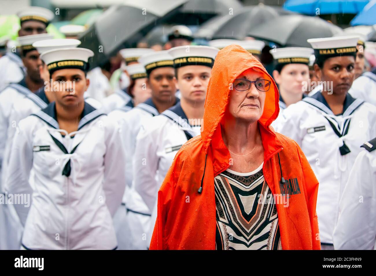 ANZAC Day als Frau in orangefarbenem Regenmantel steht mit regnernassen Marinestützern und erinnert an das Opfer und die Schrecken des Krieges in Cairns, Queensland. Stockfoto