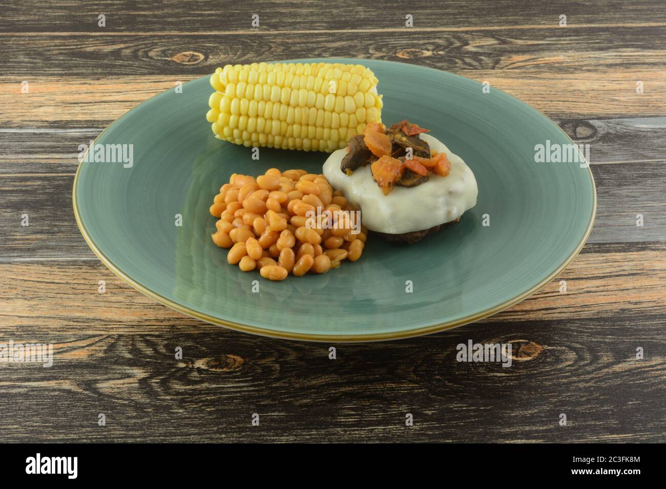 Hamburger mit geschmolzenem Provolon-Käse überzogen mit Pilzen und Tomaten, Ballen Bohnen, Mais auf dem Cob für das Essen auf blauem Teller Stockfoto