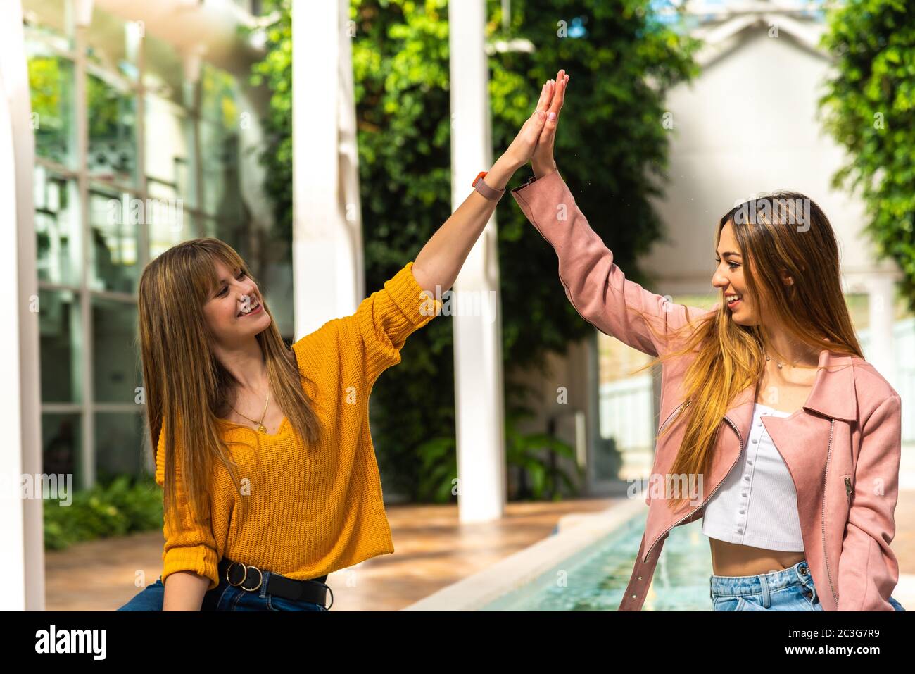 Zwei attraktive junge kaukasische Mädchen mit blonden Haaren schütteln die Hände mit ihren Armen in der Luft lächelnd an einem Brunnen in einem hell erleuchteten Raum mit dem Stockfoto