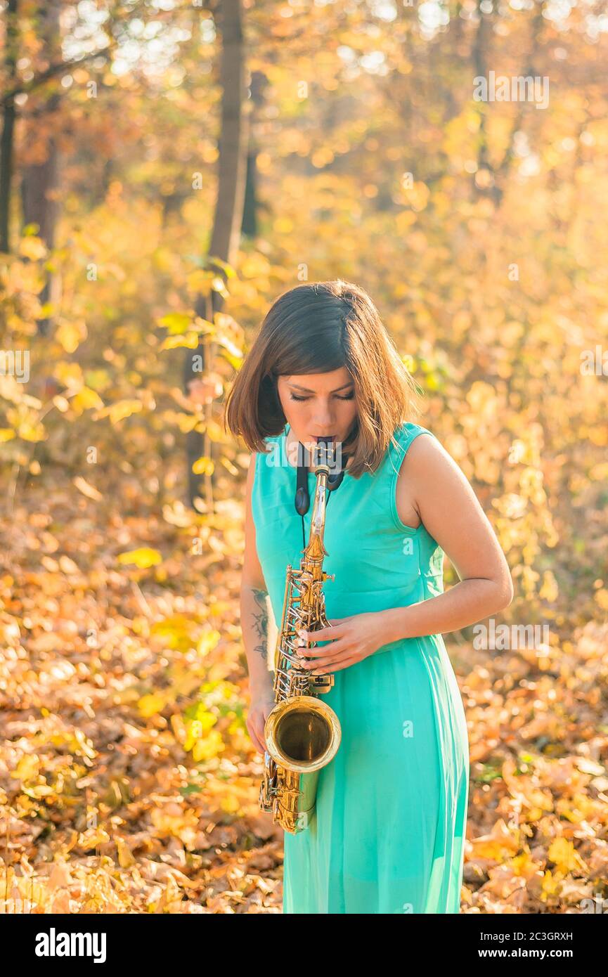 Schönes junges Mädchen mit schwarzen Haaren und einem Tattoo am Arm, in einem langen blauen Kleid gekleidet und spielt im Herbst das Altsaxophon Stockfoto